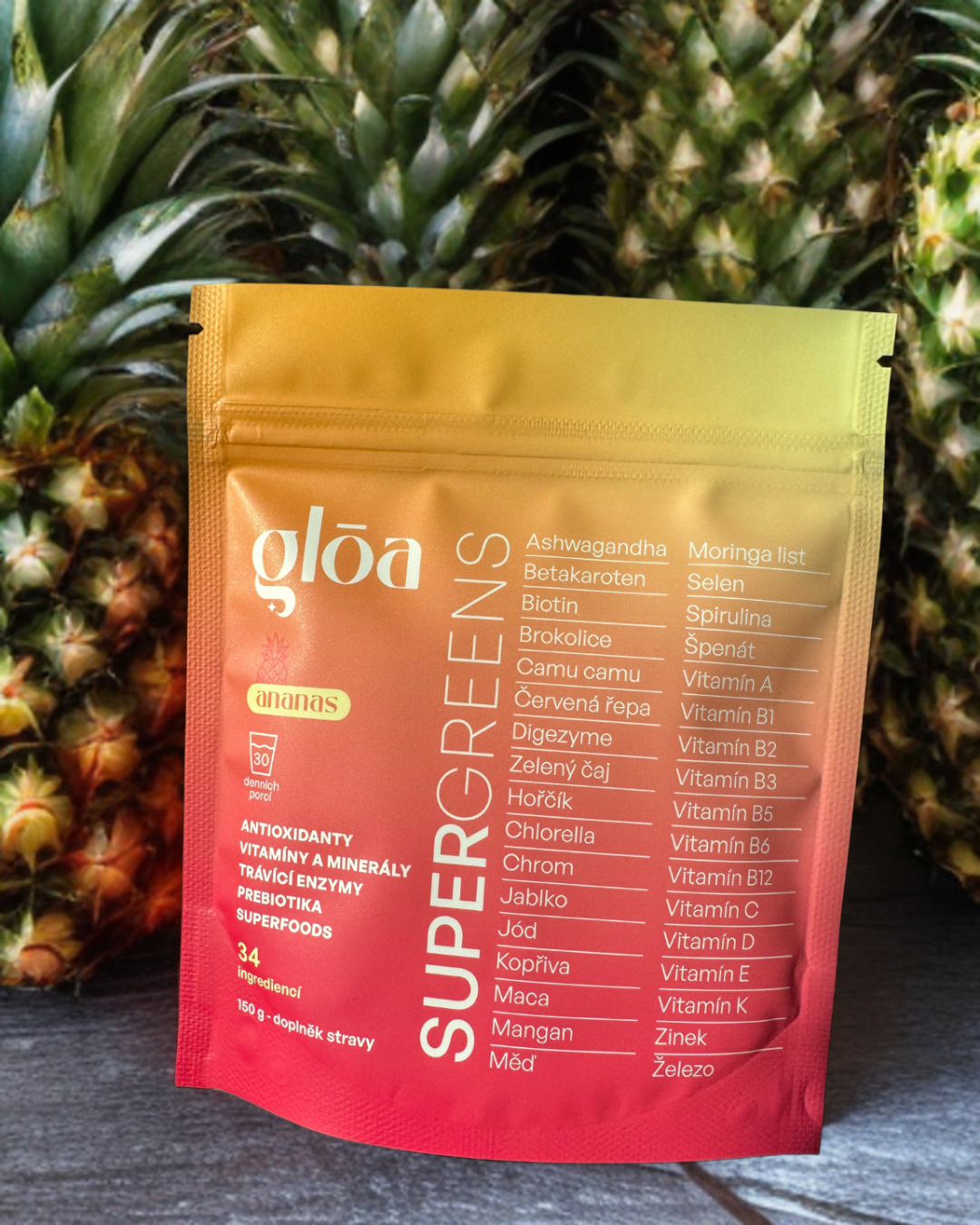 Balení Gloā Supergreens s příchutí ananasu na tropickém pozadí s čerstvými ananasy – přírodní doplněk stravy s 34 aktivními látkami pro zdraví a krásu.