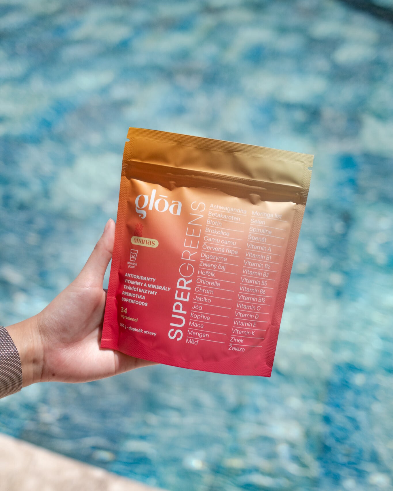 Balení Gloā Supergreens držené v ruce před modrou vodní hladinou – ideální doplněk pro zdravý start dne i na cestách.