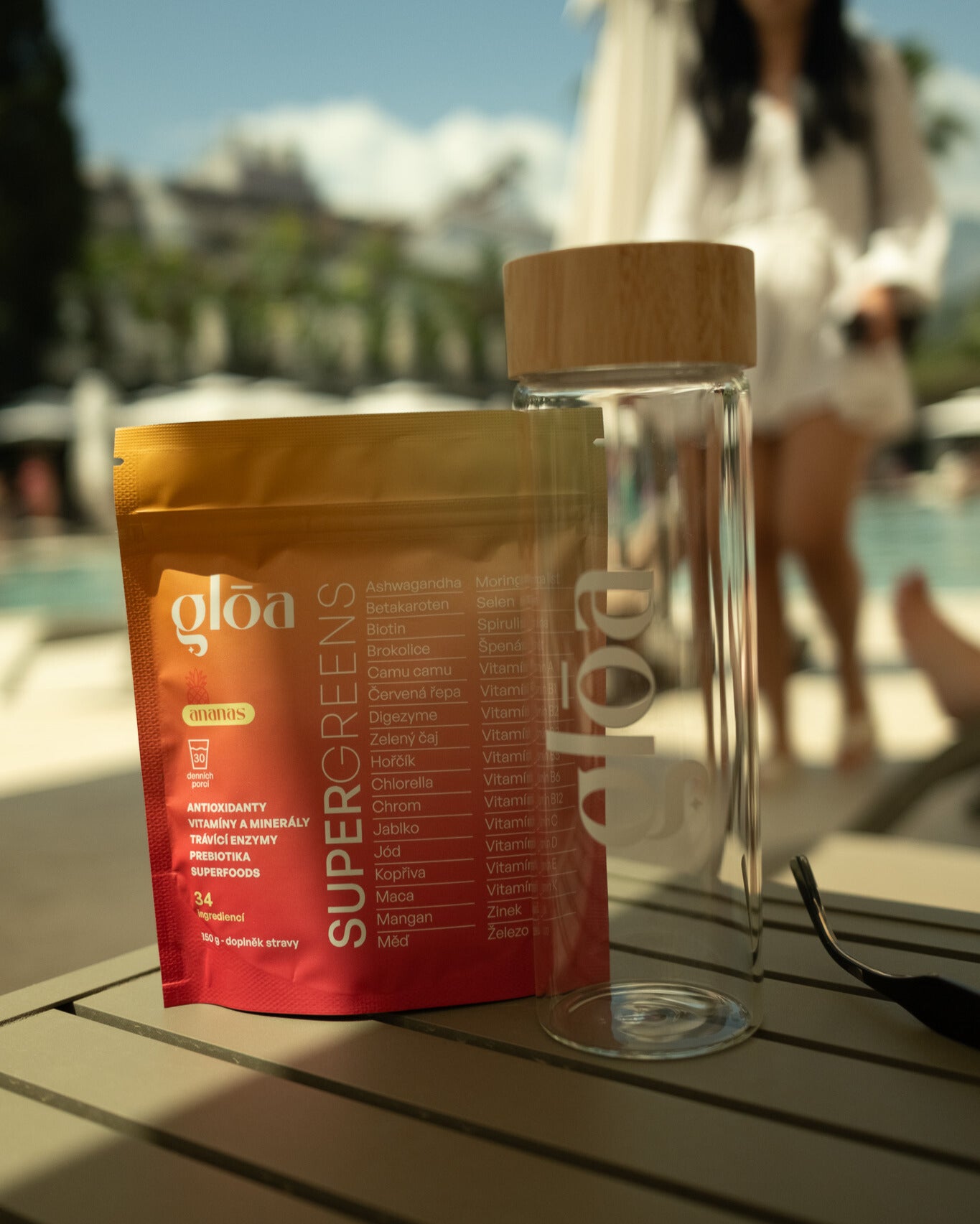 Gloā Supergreens a skleněná lahev s bambusovým víčkem na stole u bazénu s postavou v pozadí – letní lifestyle moment spojující zdraví, styl a pohodu.